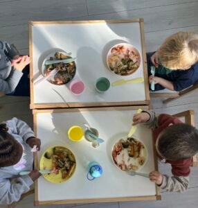 Overhead view of four children at white tables having lunch, showing colorful plates with balanced meals, child-appropriate cutlery, and varied drinking cups. The image captures the social aspect of mealtimes at our nursery.