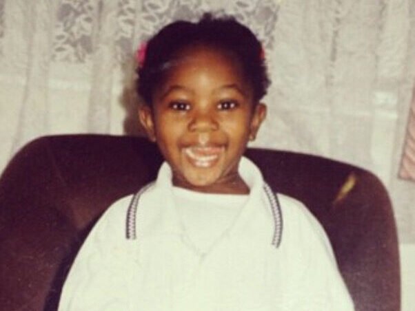 A vintage childhood photograph of Edwina in a white dress with decorative trim, displaying a bright, joyful smile. The image shows a young girl standing confidently in what appears to be a home setting with wooden furniture and lace curtains in the background.